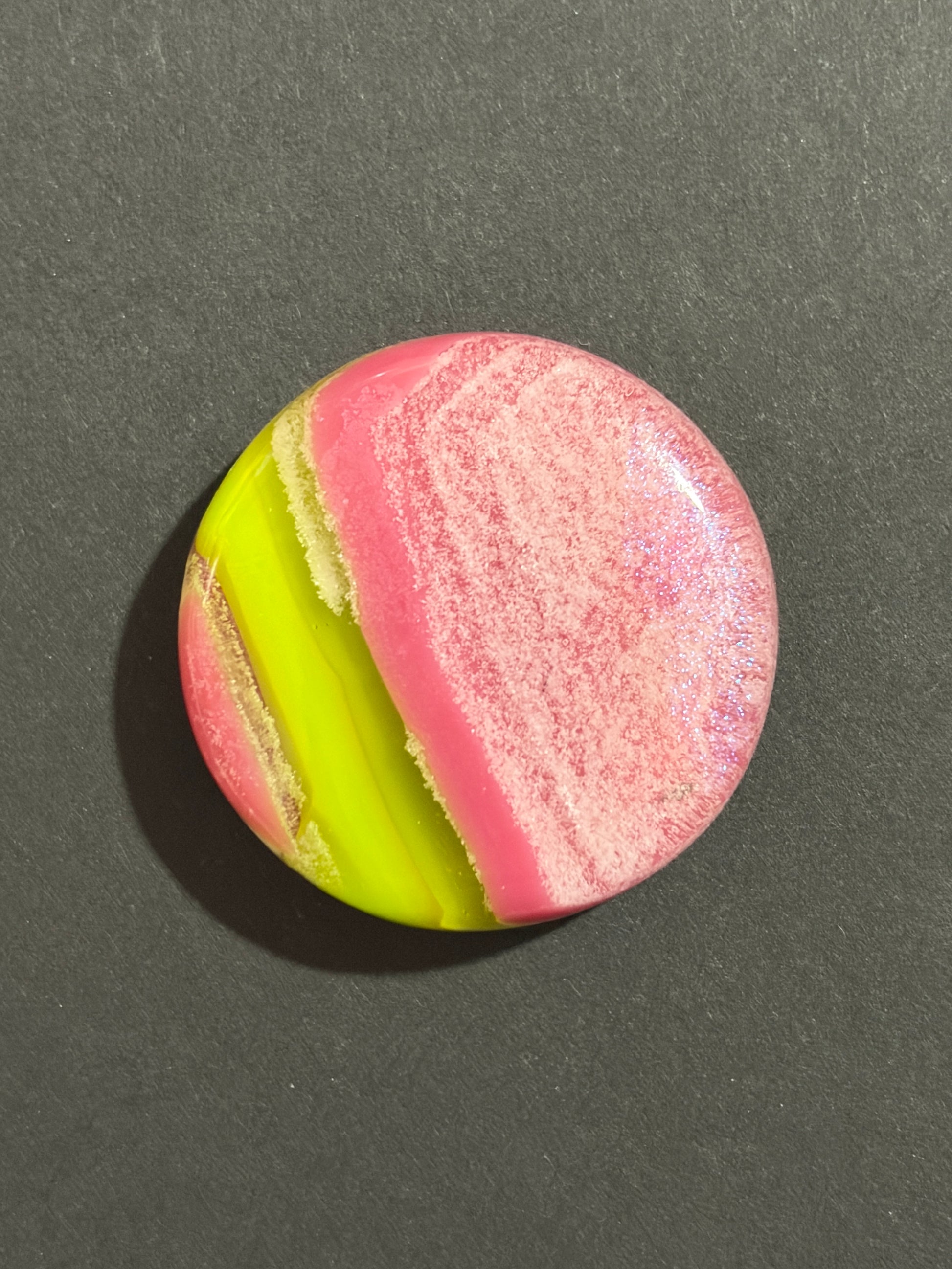 A round cabochon gemstone featuring a marbled pattern of bright pink, neon yellow-green, and cream swirls, displayed on a dark gray textured background.