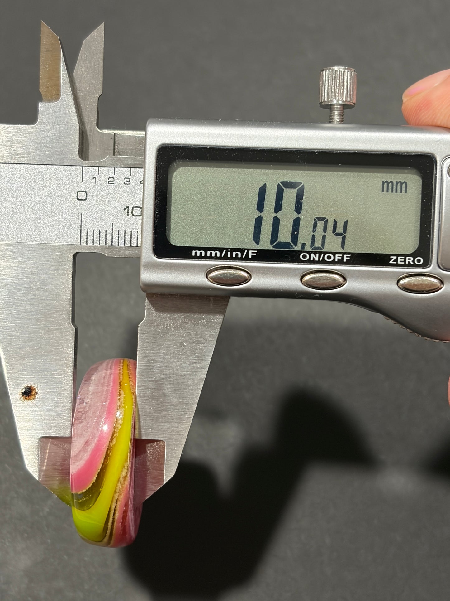 A close-up of a digital caliper measuring a small oval ring or bead. The caliper display reads 10.