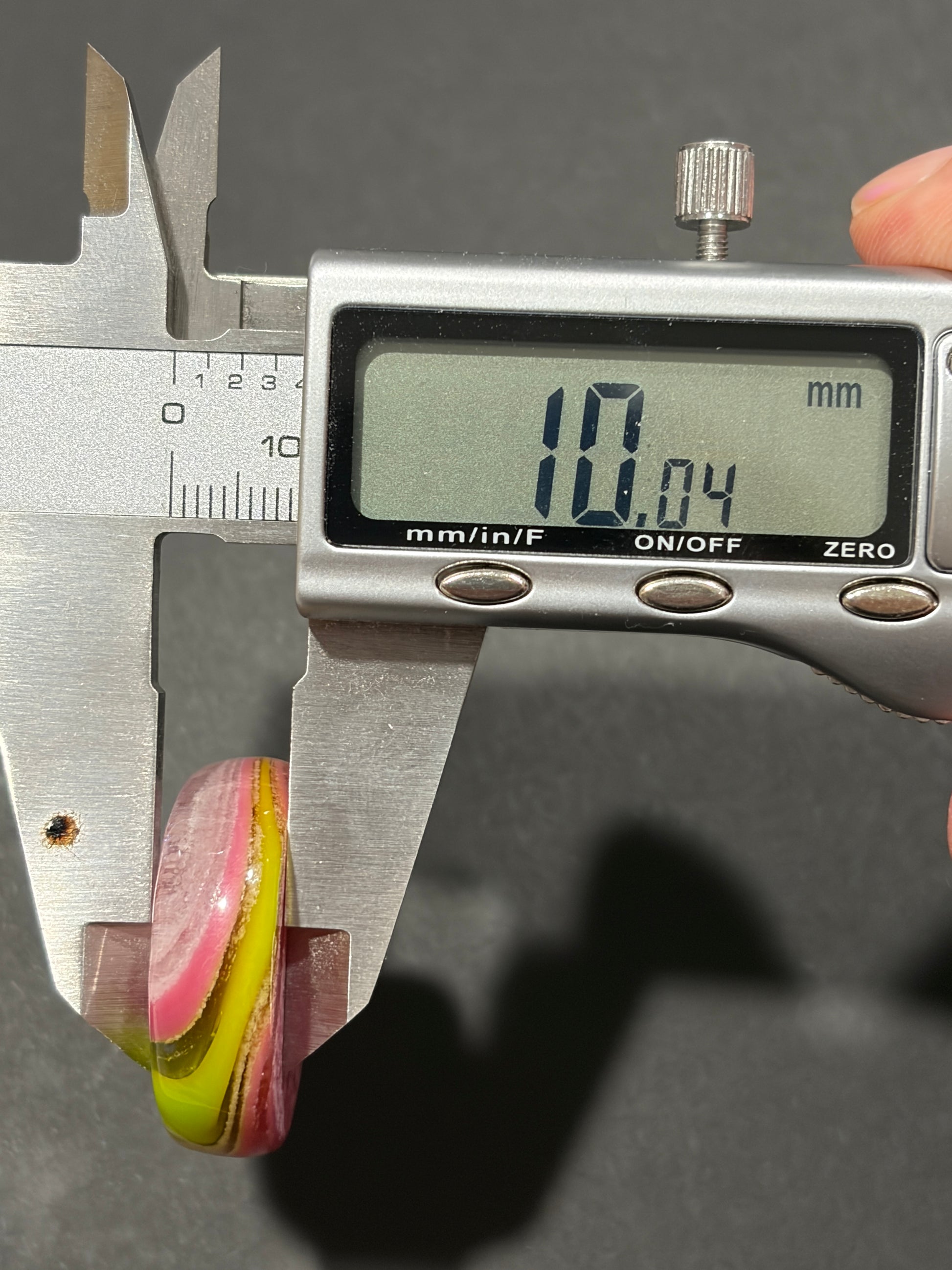 A close-up of a digital caliper measuring a small oval ring or bead. The caliper display reads 10.